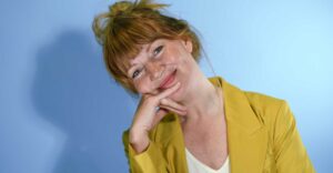 Portret van een vrouw met rood haar in een gele blazer, tegen een blauwe studioachtergrond.